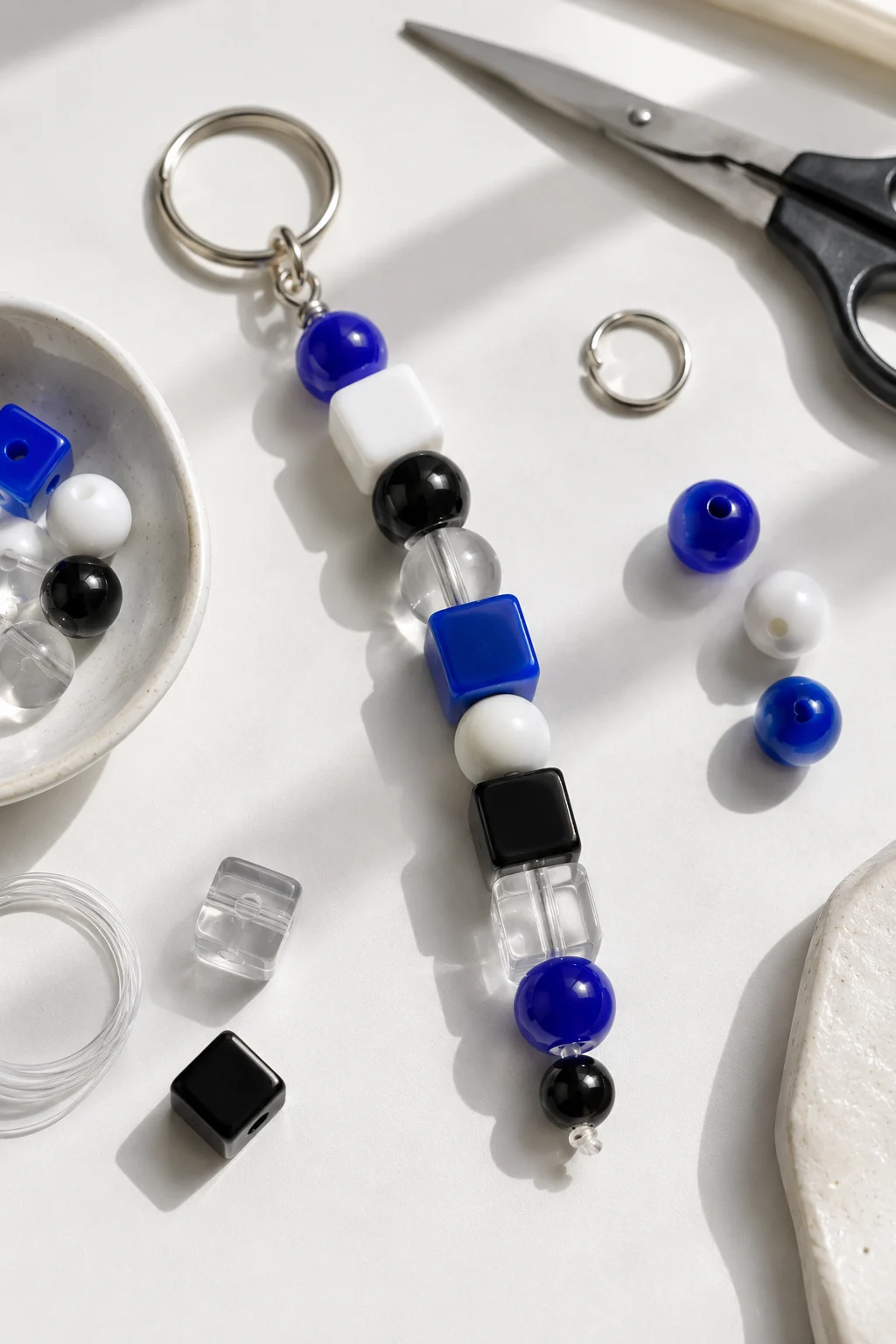 A finished color block bead keychain featuring cobalt blue, white, black, and clear round and square beads arranged in a modern alternating pattern, securely threaded onto clear cord and attached to a silver metal keyring, with extra beads, scissors, and craft supplies visible around the handmade keychain on a bright tabletop.