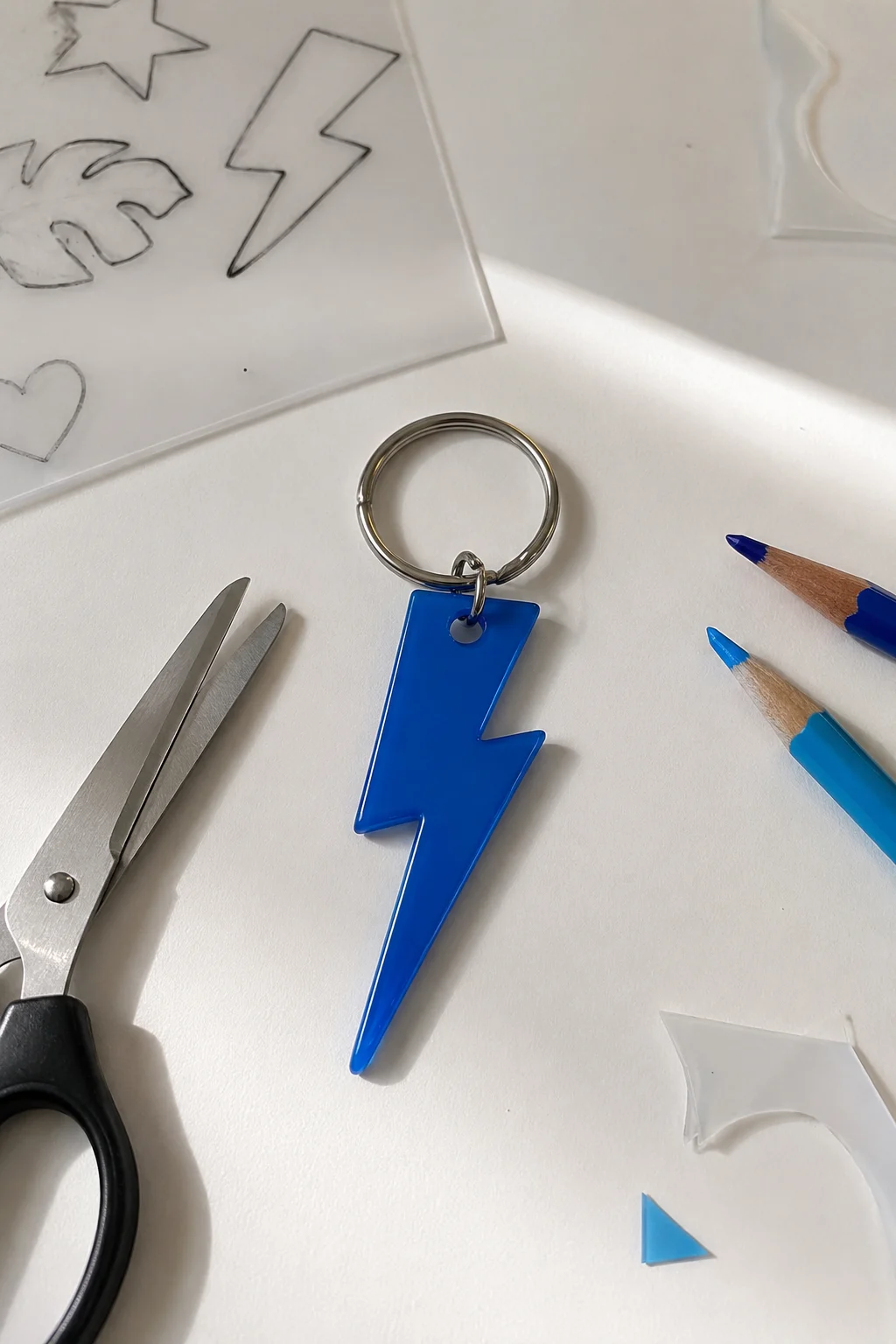 A finished bright blue lightning bolt keychain made from shrink plastic, featuring a clean angular graphic silhouette with smooth edges and a polished surface, attached through a small connector hole to a silver metal keyring, with blue pencils, scissors, and hand-drawn shape templates visible around the DIY project.