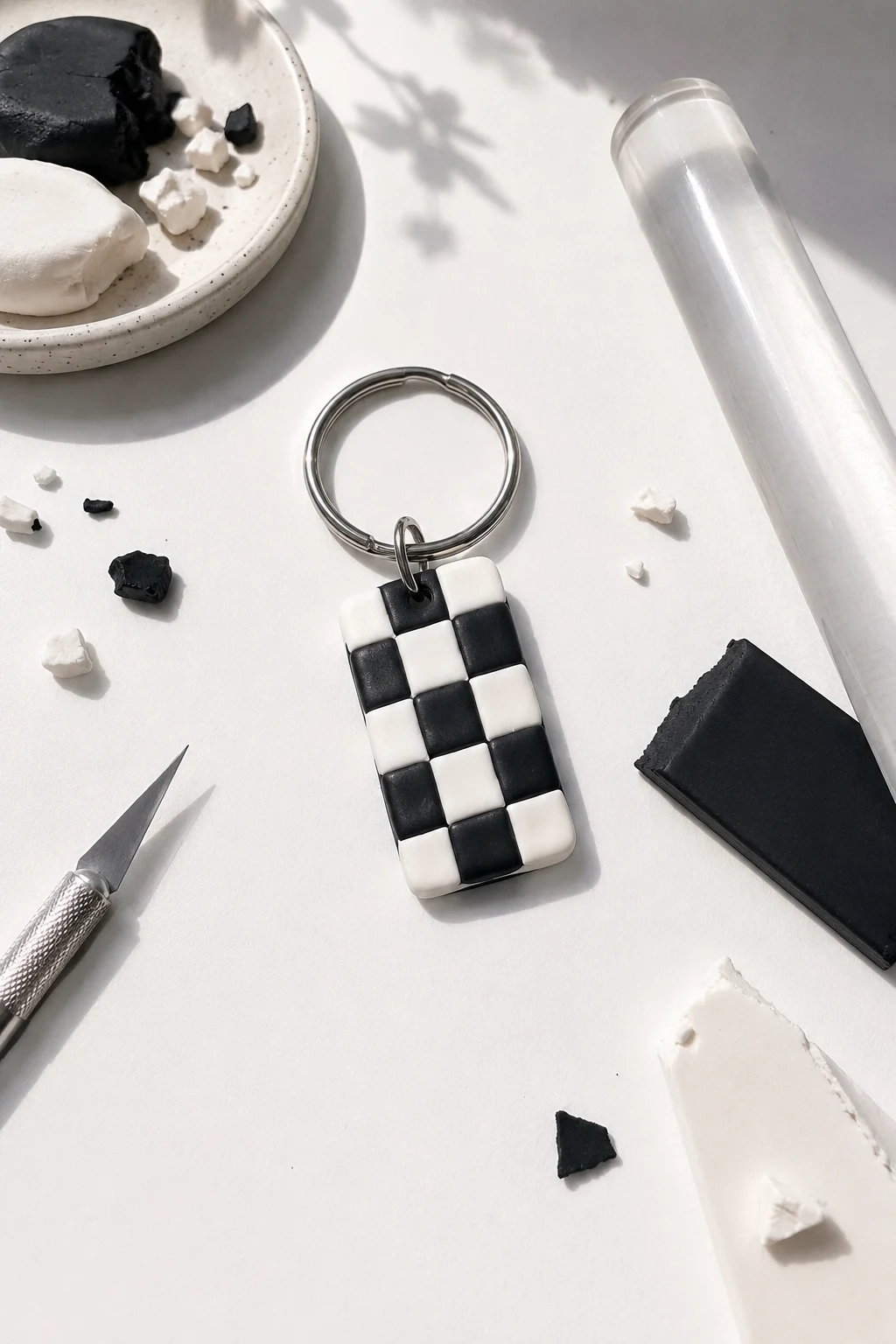 A finished rectangular polymer clay keychain featuring an evenly spaced black and white checkerboard pattern with rounded corners, attached to a polished silver metal keyring, surrounded by black and white clay pieces, a rolling tool, and small clay scraps on a bright crafting workspace.