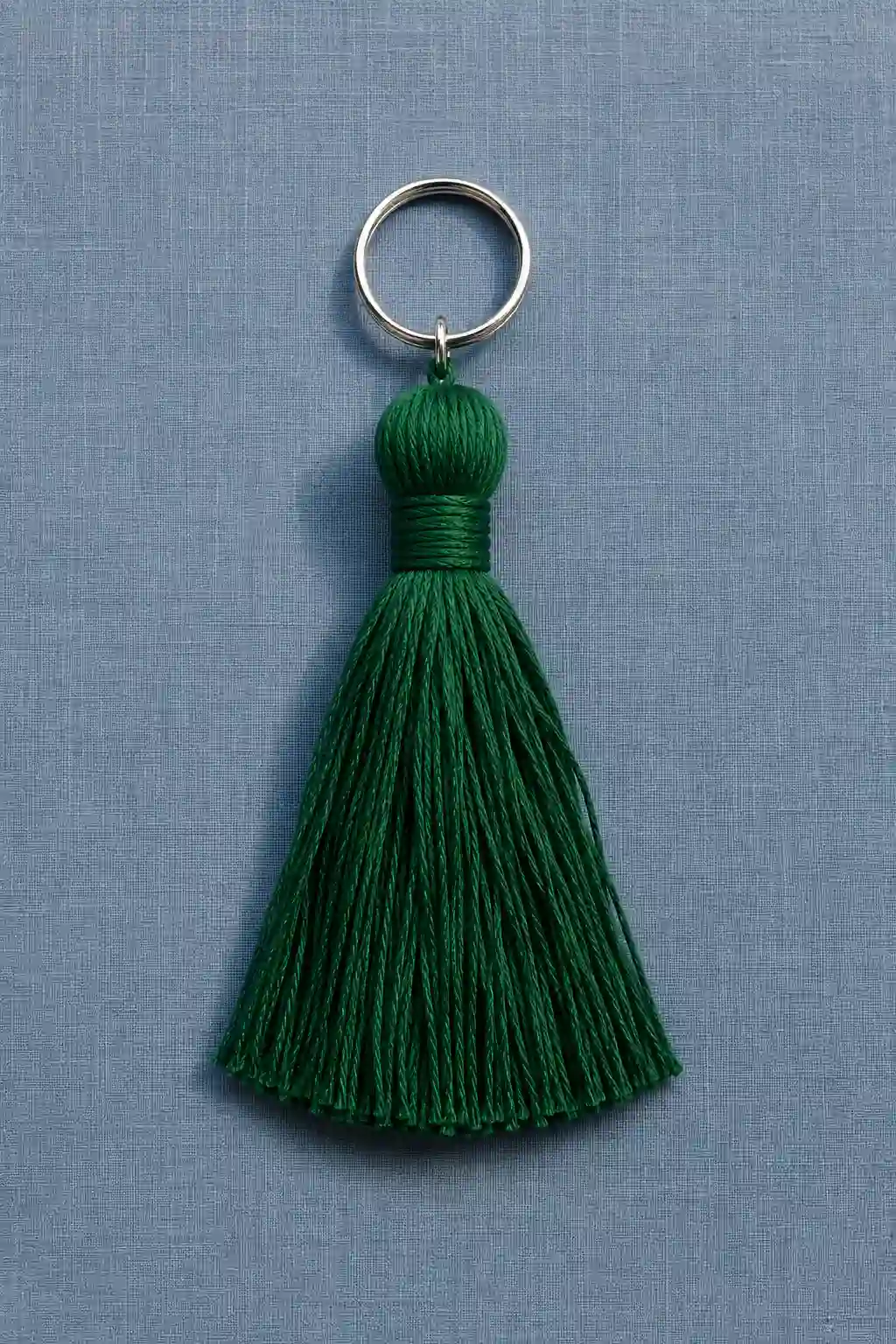 A finished deep emerald green tassel keychain made from multiple strands of smooth embroidery thread, featuring a rounded wrapped top, tightly secured center band, evenly trimmed flowing tassel strands, and a small silver metal keyring, photographed against a soft blue textured background.