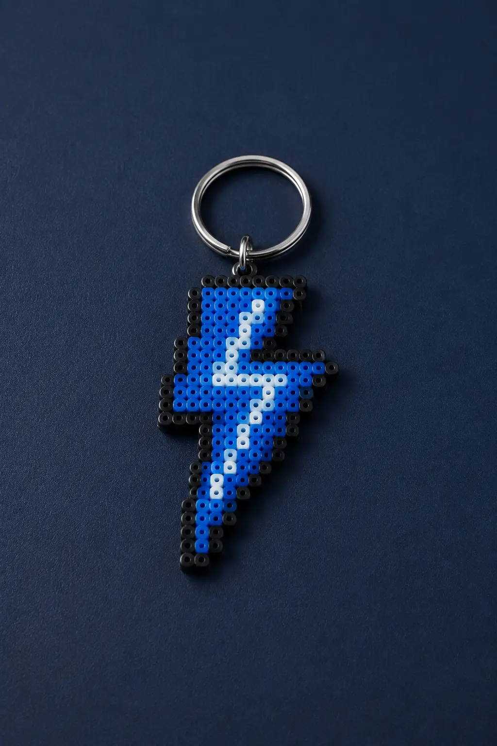 A finished pixel-style lightning bolt keychain created from fused blue, white, and black fuse beads, arranged in a bold geometric pattern with a dark outline and attached to a silver metal keyring, photographed closely against a deep navy textured surface to emphasize the colorful pixel details.