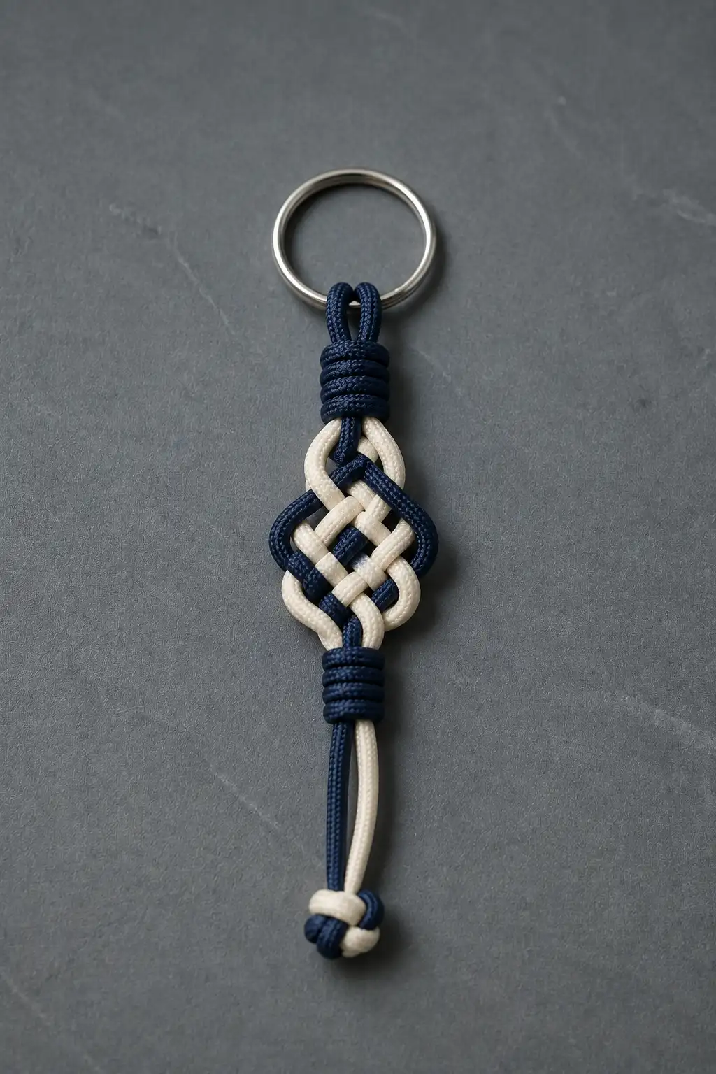 A finished two-tone paracord keychain featuring an intricate woven geometric knot pattern made from navy blue and cream paracord, with tightly wrapped sections at the top and bottom and a polished silver metal keyring, photographed on a dark gray textured surface to highlight the detailed handmade weave.