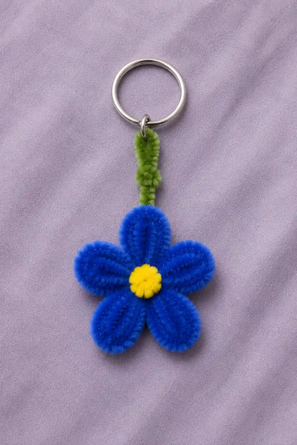 A finished pipe cleaner flower keychain featuring five rounded royal-blue chenille petals, a small bright yellow textured center, and a short green chenille stem connected to a silver metal keyring, photographed against a soft lavender background to clearly show the complete handmade flower shape and fluffy texture.