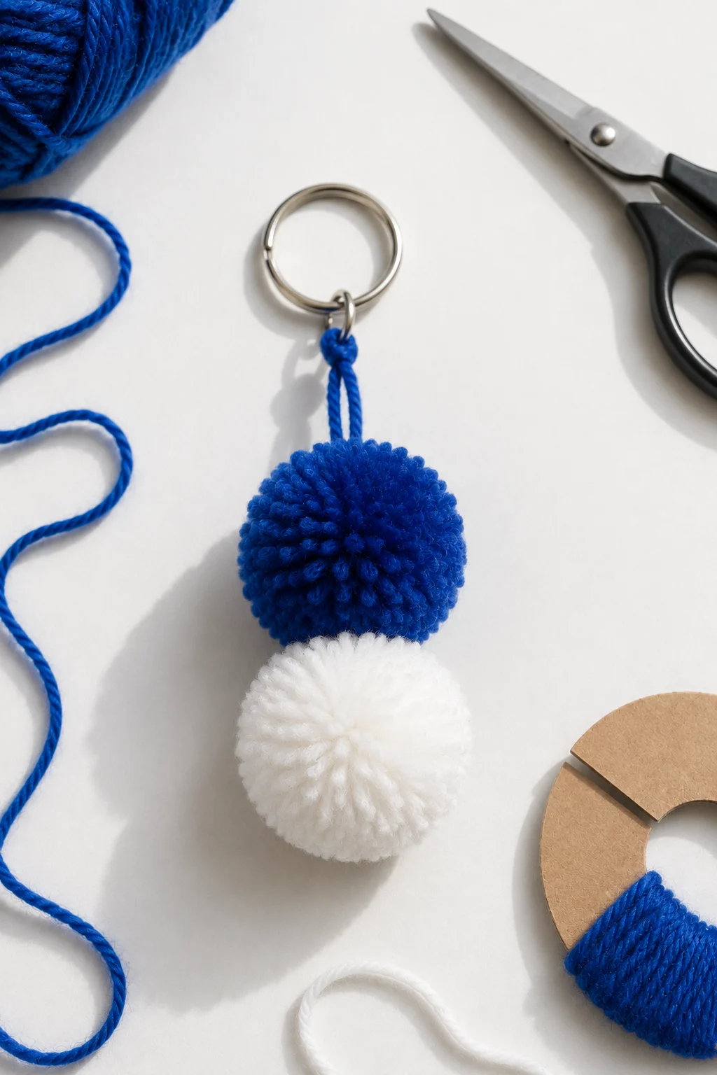 A finished two-tone pom-pom keychain made with soft royal blue and white yarn, featuring two fluffy round pom-poms attached vertically to a silver metal keyring, with blue yarn, scissors, and a cardboard pom-pom template arranged nearby during the DIY crafting process.