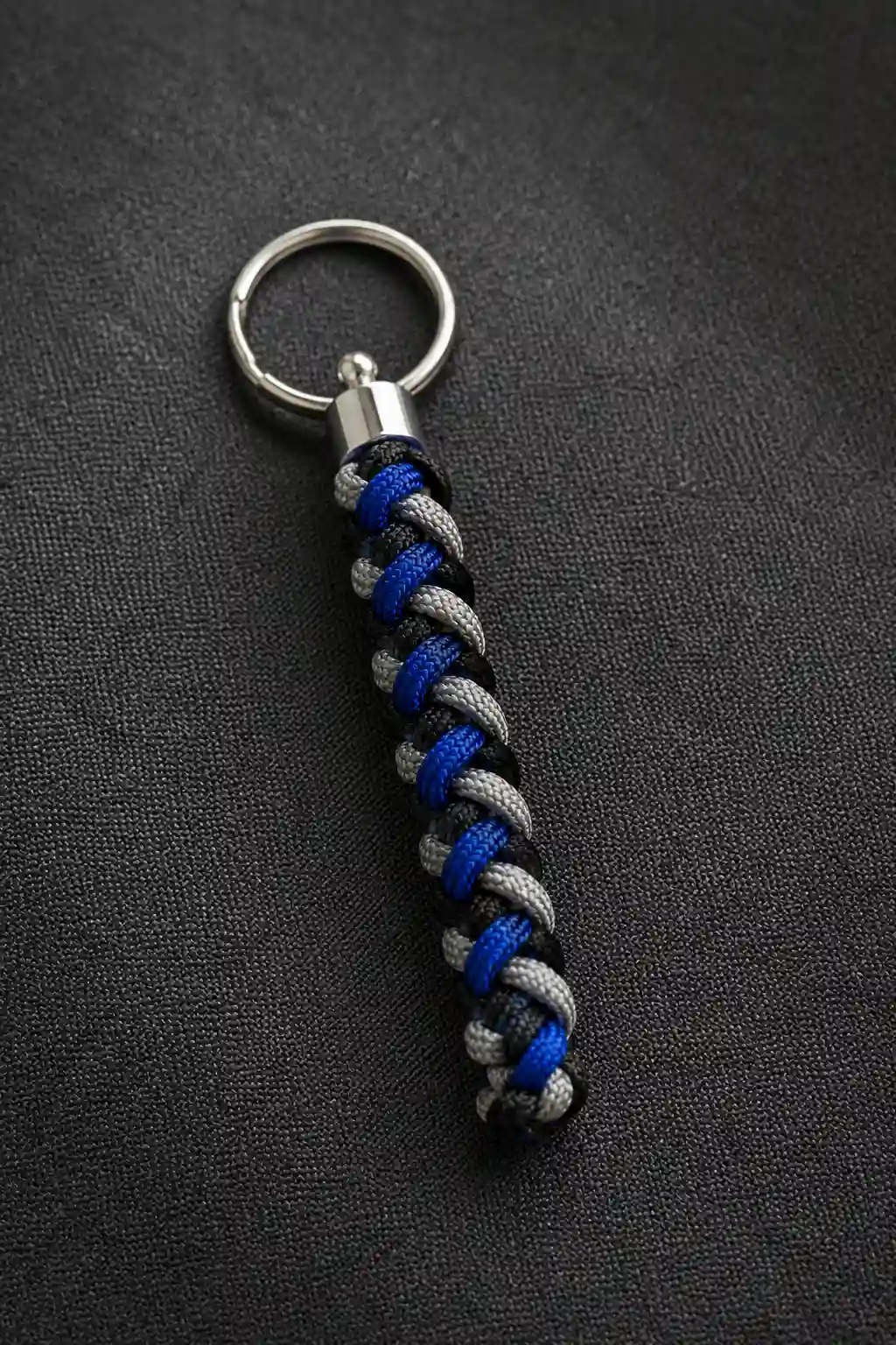 A finished braided cord keychain featuring tightly woven cobalt blue, black, and silver-gray cords in a long geometric braid, attached to a polished silver metal keyring with a secure metal connector, photographed closely on a dark textured surface to highlight the detailed handmade pattern.