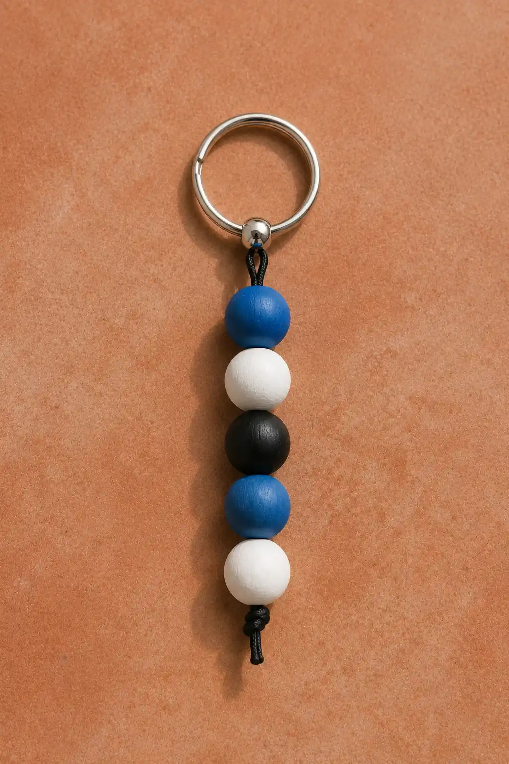 A finished painted wooden bead keychain featuring smooth round beads in cobalt blue, white, and matte black arranged in an alternating color block pattern, threaded onto black cord and attached to a silver metal keyring, photographed against a warm tan craft surface with soft natural shadows.