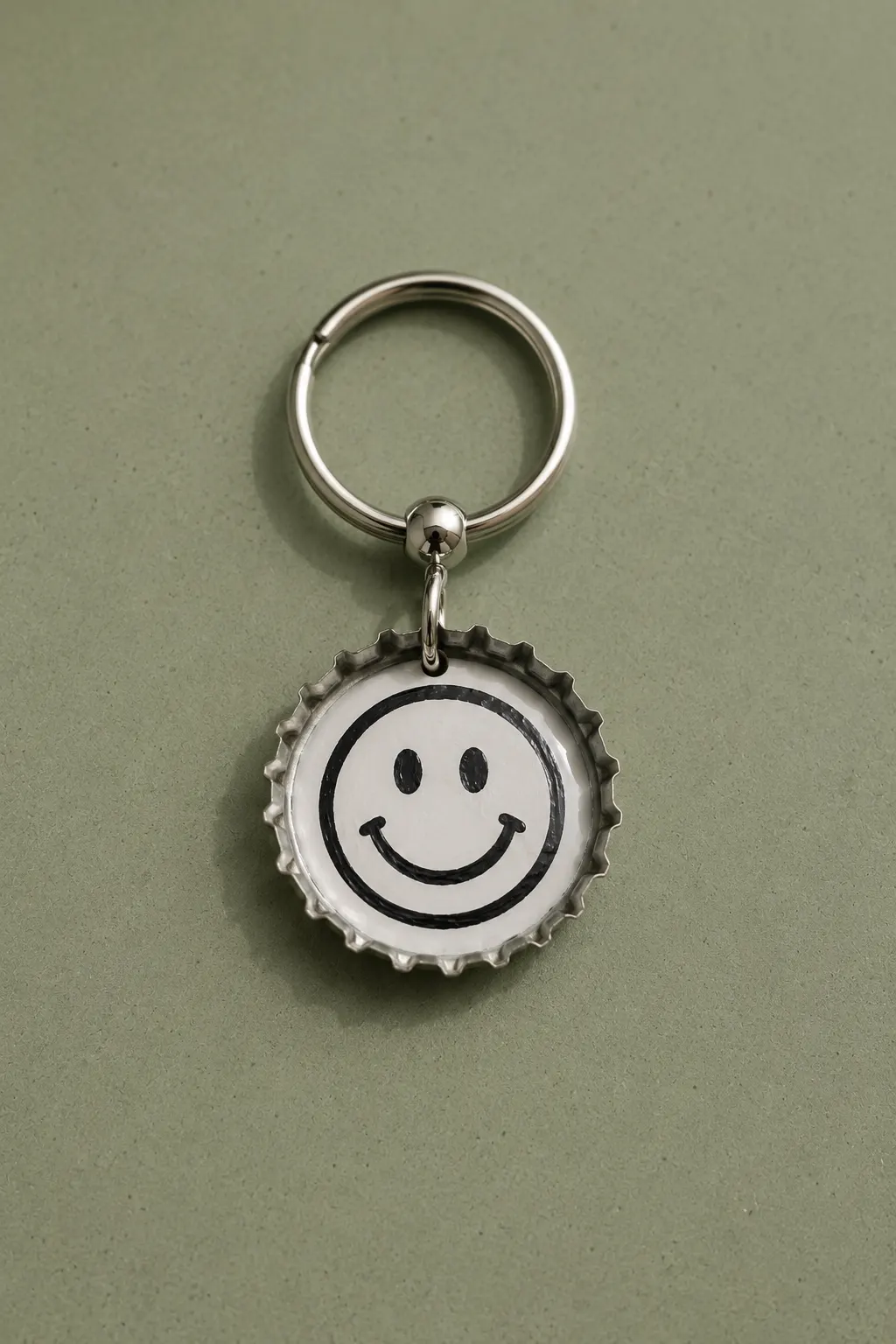 A finished recycled bottle cap keychain featuring a silver metal bottle cap transformed into a playful graphic charm with a bold black smiley face on a white center, attached to a polished silver keyring and photographed on a muted green surface with a clean modern handmade aesthetic.