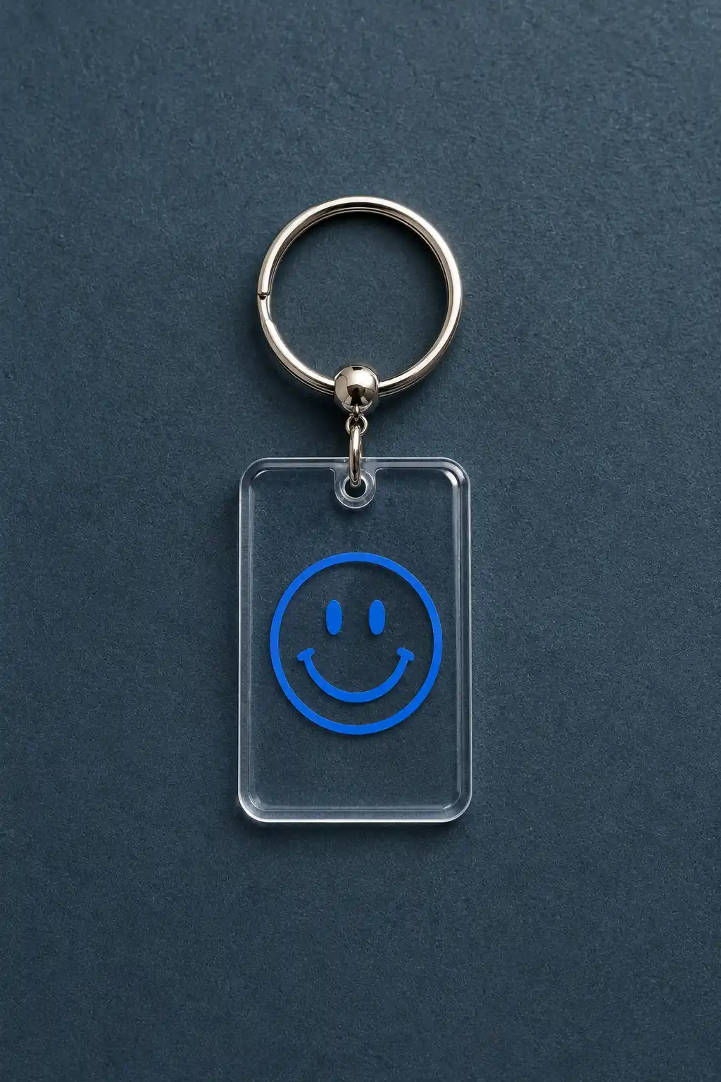 A finished clear rectangular graphic keychain made from transparent plastic, featuring a bold electric-blue smiley face graphic in the center and a small connector hole at the top, attached to a polished silver metal keyring and photographed on a dark navy textured surface for a modern minimalist look.
